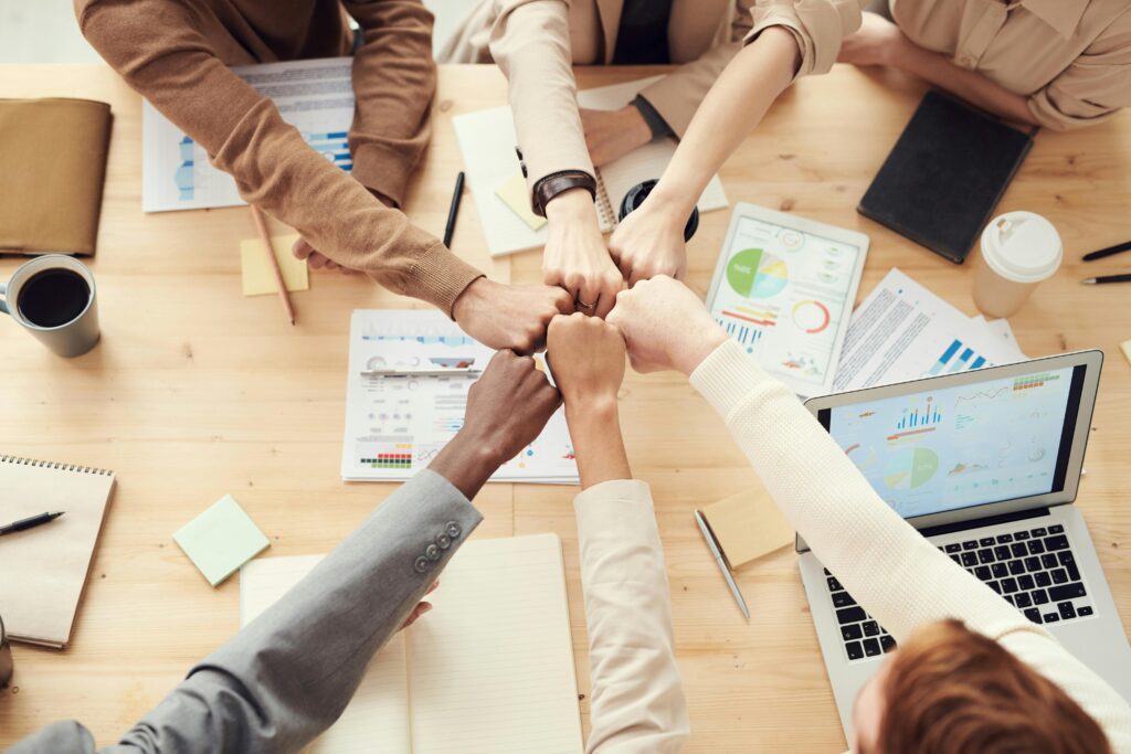 pexels-photo-3184438-3184438 Top view of a diverse team fist bumping over a meeting table with paperwork and laptops, symbolizing teamwork.